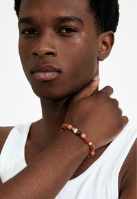 Pulsera de cuentas con piedras rojas, naranjas y marrones de texturas suaves, llevada en una muñeca sobre un fondo claro.