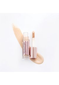 Corrector en un tubo transparente con tapa de color rosa dorado, que cuenta con un aplicador tipo doe-foot. Acompañado de una muestra del producto en un tono beige claro.