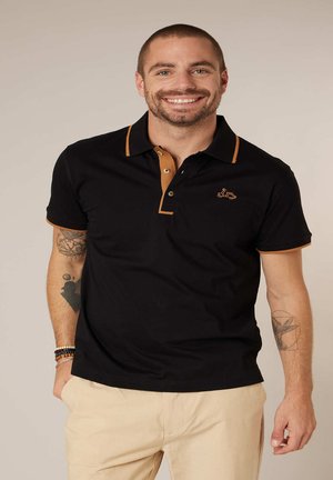 Homme souriant aux cheveux courts portant un polo noir avec des bords marron et un pantalon beige clair, debout les mains dans les poches.