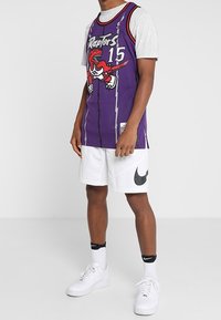 Man i en lila Toronto Raptors #15 baskettröja, vita Nike-shorts, vita Nike-strumpor och vita Nike-sneakers som står mot en enfärgad bakgrund.