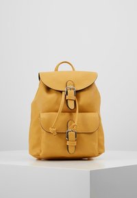 Malý hořčičně žlutý kožený batoh s přední kapsou a stříbrnými sponami, stojící na bílé platformě na jednobarevném pozadí.