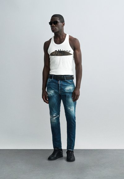 Uomo con occhiali da sole neri, canotta bianca con grafica, jeans blu strappati, cintura nera e scarpe da ginnastica nere in piedi contro una parete grigia uniforme.