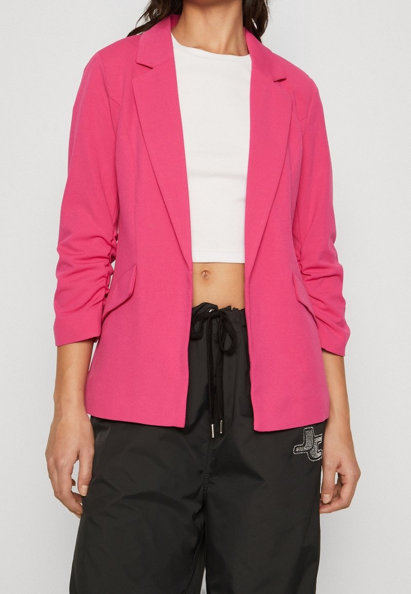 Femme portant un blazer rose vif par-dessus un crop top blanc et un pantalon noir à cordon avec un petit logo brodé.