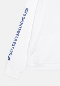 Bílá mikina s kapucí, na které je na rukávu v modré barvě vytištěno "Nike Sportswear Est. 1972". Obsahuje přední kapsu a žebrované manžety.