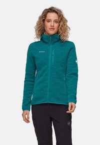 Giacca in pile turchese con colletto alto, zip integrale e due tasche con zip. Presenta una texture sottile e il logo del marchio sul petto e sulla manica.