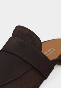 Chaussure à enfiler en daim brun foncé avec une large bride et une semelle intérieure couleur fauve sur un fond clair uni.
