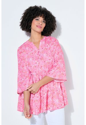 Kvinde med krøllet hår iført en lyserød blomstret bluse med 3/4-ærmer og hvide bukser, stående mod en lys baggrund.