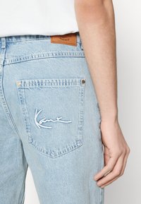 Jeans de denim azul claro con un bolsillo trasero que presenta un logotipo bordado en blanco que dice "Kane", remaches de bronce y una etiqueta de cuero marrón en la cinturilla.