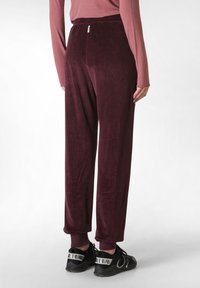 Bordeaux fluwelen broek met een ontspannen pasvorm, elastische tailleband en ribgebreide enkels, gecombineerd met zwarte sneakers met witte accenten.
