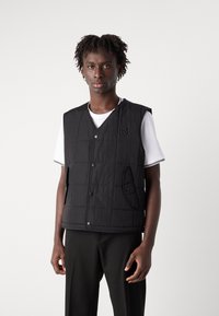 Vest negru matlasat cu decolteu în V, închidere cu nasturi și buzunar lateral. Purtați peste un tricou alb cu bordură neagră, expus pe un fundal simplu.