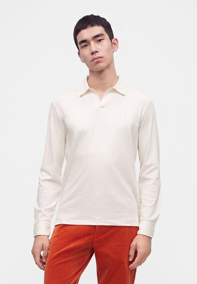 Νέος άνδρας που φοράει ένα μακρυμάνικο off-white πόλο και πορτοκαλί παντελόνι από κοτλέ, στέκεται μπροστά σε απλό λευκό φόντο.