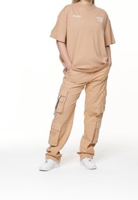 Béžové oversized tričko s nápisem "PGDR" a "NEVER TOO LATE", doplněné béžovými cargo kalhotami s několika kapsami a bílými teniskami.