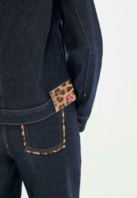 Veste en denim foncé avec poche à rabat en imprimé léopard contrastant et logo rose. Présente des coutures apparentes et un tissu texturé.