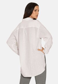 Weißes, langärmeliges Hemd aus leichtem, strukturiertem Stoff, mit Rückenpasse und verlängertem Saum, kombiniert mit karierter Hose.