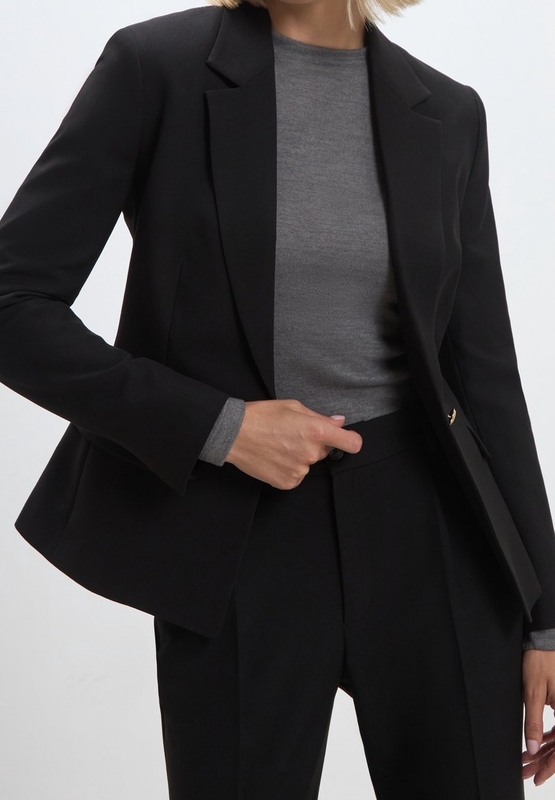 Femme ajustant un bouton sur un blazer noir porté sur un haut gris à manches longues, associé à un pantalon noir sur mesure, sur un fond clair uni.