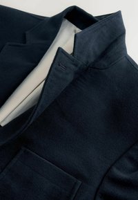 Giacca in misto lana blu navy con superficie testurizzata, dotata di un colletto piccolo e una tasca sul petto. Foderata con un tessuto di colore chiaro in contrasto.