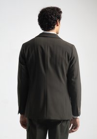 Hombre con cabello rizado, vistiendo un blazer oscuro y pantalones a juego, de pie de espaldas contra un fondo blanco liso.