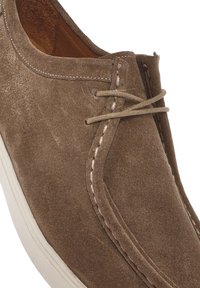 Scarpa in suede marrone con una consistenza morbida, punta arrotondata, dettagli in cucitura bianca e un rivestimento in pelle marrone, con due fori per i lacci e i lacci stessi.