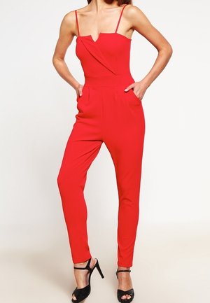 Kvinde iført en ærmeløs rød jumpsuit med tynde stropper og sorte høje hæle med åbne tæer, hænderne i lommerne.