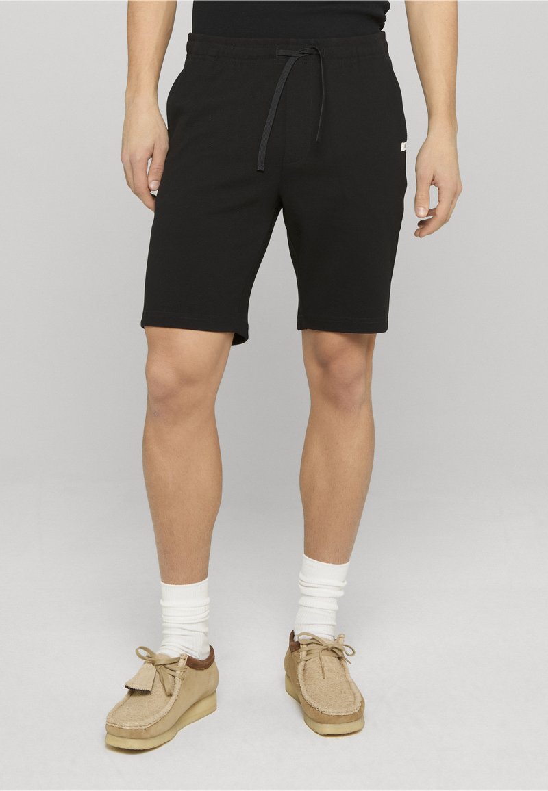 Shorts en coton noir avec une taille ajustable à cordon, poches latérales et une texture lisse. Portés avec des chaussures beige et des chaussettes blanches.