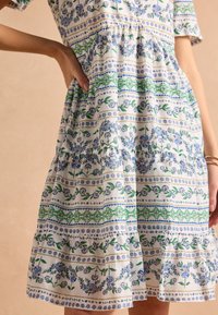 Bavlnené kvetinové šaty s krátkymi rukávmi, volánovou sukňou a mnohobarevnými kvetinovými vzormi v modrej a zelenej na bielom pozadí.