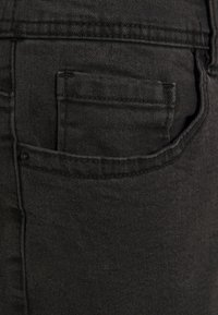 Jeans de mezclilla negra con un solo bolsillo trasero, presentando una tela texturizada y sutilezas de costura a lo largo de las costuras.