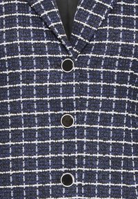 Veste en tweed bleue et noire avec motif à carreaux blancs et trois grands boutons noirs à bordure argentée sur le devant.
