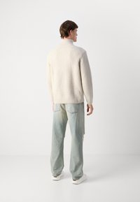 Beige Ripp-Strickpullover mit hohem Kragen, kombiniert mit verblassten hellblauen weiten Jeans und weißen Sneakers, von hinten betrachtet.