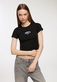 Maglietta nera cropped con maniche corte e lavorazione a maglia testurizzata, con la scritta bianca "BROOKLYN NYC" sul petto. Abbinata a jeans grigi.