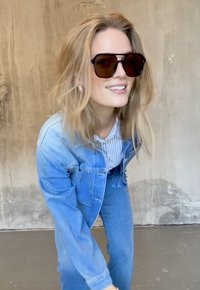 Zonnebril met donkere glazen, oversized vierkante vorm. Model draagt een lichtblauwe denimjas over een gestreept blouse. Informele setting.