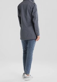 Dlouhý šedý kabát z texturované látky, s vysokým límcem a volným střihem, v kombinaci s modrými džíny a šedými teniskami.