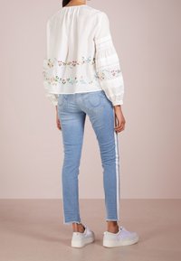 Blusa blanca bordada con patrones florales, mangas amplias, combinada con jeans azul claro con rayas laterales y zapatillas blancas.