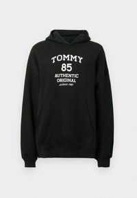 Čierna mikina s kapucňou z mäkkej látky s klokanie vrecko. Na prednej strane je biely nápis: „TOMMY 85 AUTHENTIC ORIGINAL since 1985".