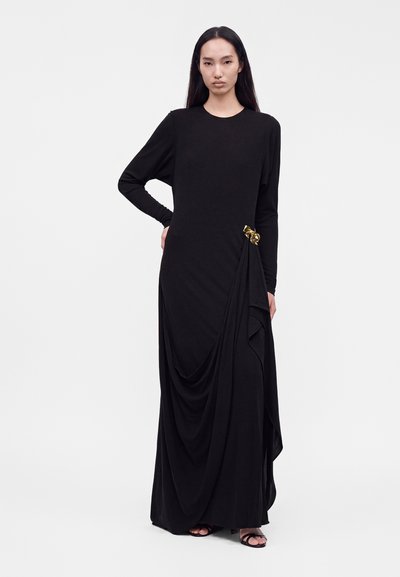 Femme aux longs cheveux noirs portant une longue robe noire avec un tissu drapé et une broche dorée, debout, une main sur la hanche.