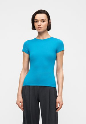 Γυναίκα με κοντά σκούρα μαλλιά που φοράει εφαρμοστή φωτεινή μπλε κοντομάνικη μπλούζα και φαρδιά σκούρα γκρι παντελόνια, στέκεται μπροστά σε απλό φόντο.