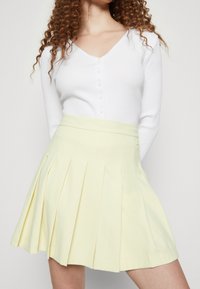 Bílý žebrovaný top s dlouhým rukávem a knoflíky spárovaný se žlutou plisovanou sukní, která má přiléhavý pas a měkkou texturu látky.