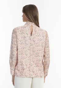 Blomstret bluse i lys pink med metalliske accenter. Har en høj halsudskæring, nøglehul bagpå og lange ærmer med sammensnørrede manchetter.