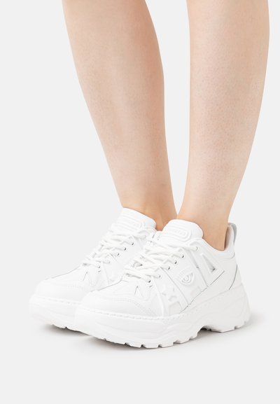 Baltas apavi ar masīvu zoli, saistīšanas dizains, dekoratīvi akcenti un teksturēta virsma ar zvaigznēm un acs rakstiem.