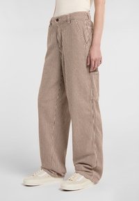 Weite Hosen mit braunen und weißen vertikalen Streifen, Knopfverschluss und seitlichen Taschen. Kombiniert mit cremefarbenen Sneakers. Strukturierter Stoff.