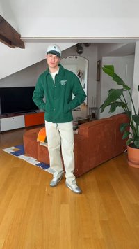 Νέος άνδρας με πράσινο μπουφάν, λευκό καπέλο και μπεζ παντελόνι στέκεται σε ένα μοντέρνο σαλόνι με ξύλινο πάτωμα, πορτοκαλί καναπέ, τηλεόραση και γλάστρα με φυτό.