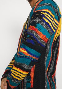 Monivärinen neulecardigan, jossa on vaakaraitoja sinisen, oranssin, keltaisen ja mustan sävyissä. Paksut tekstuurit, joissa on erilaisia kuvioita ympäriinsä.