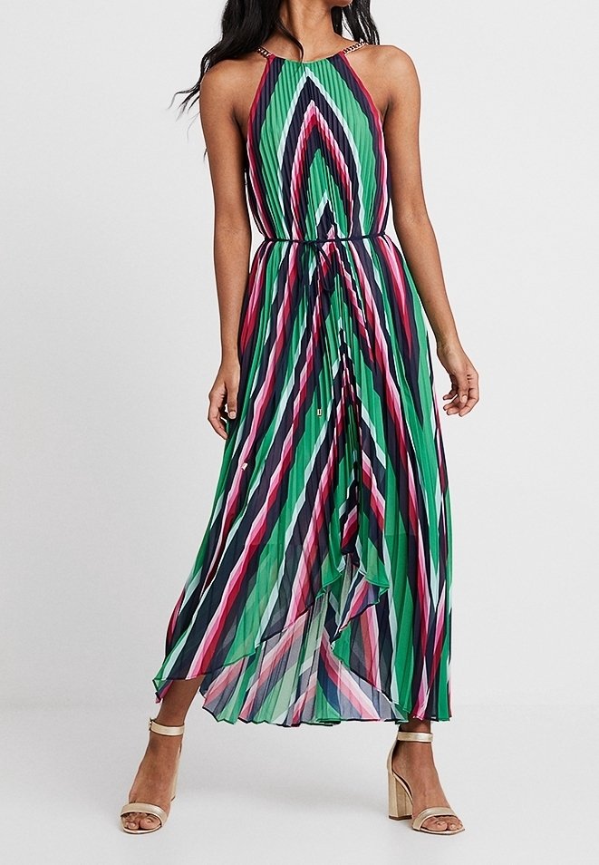 Femme portant une robe longue plissée sans manches à rayures diagonales vertes, roses, noires et blanches, assortie à des sandales à talons hauts beiges.