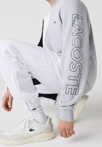 Šedá mikina s černými detaily a textem "LACOSTE" na rukávu; doplněná o bílé sportovní kalhoty a bílé tenisky.