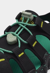 Μαύρο και πετρόλ σανδάλι με ρυθμιζόμενα ελαστικά κορδόνια τιρκουάζ που ασφαλίζουν με γκρι κλιπ και μαύρα λουριά με κίτρινη ραφή.