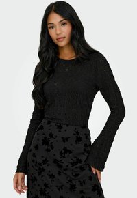 Donna con lunghi capelli neri, che indossa un top a maniche lunghe in pizzo nero e una gonna nera con fantasia floreale, in piedi di fronte a uno sfondo neutro.