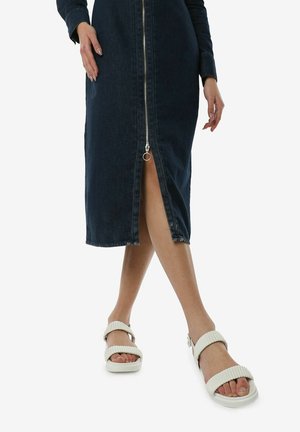 Robe midi en denim de couleur bleu foncé avec une fermeture éclair à l'avant, des manches longues et une fente à l'avant, associée à des sandales blanches à côtes avec semelles plates.
