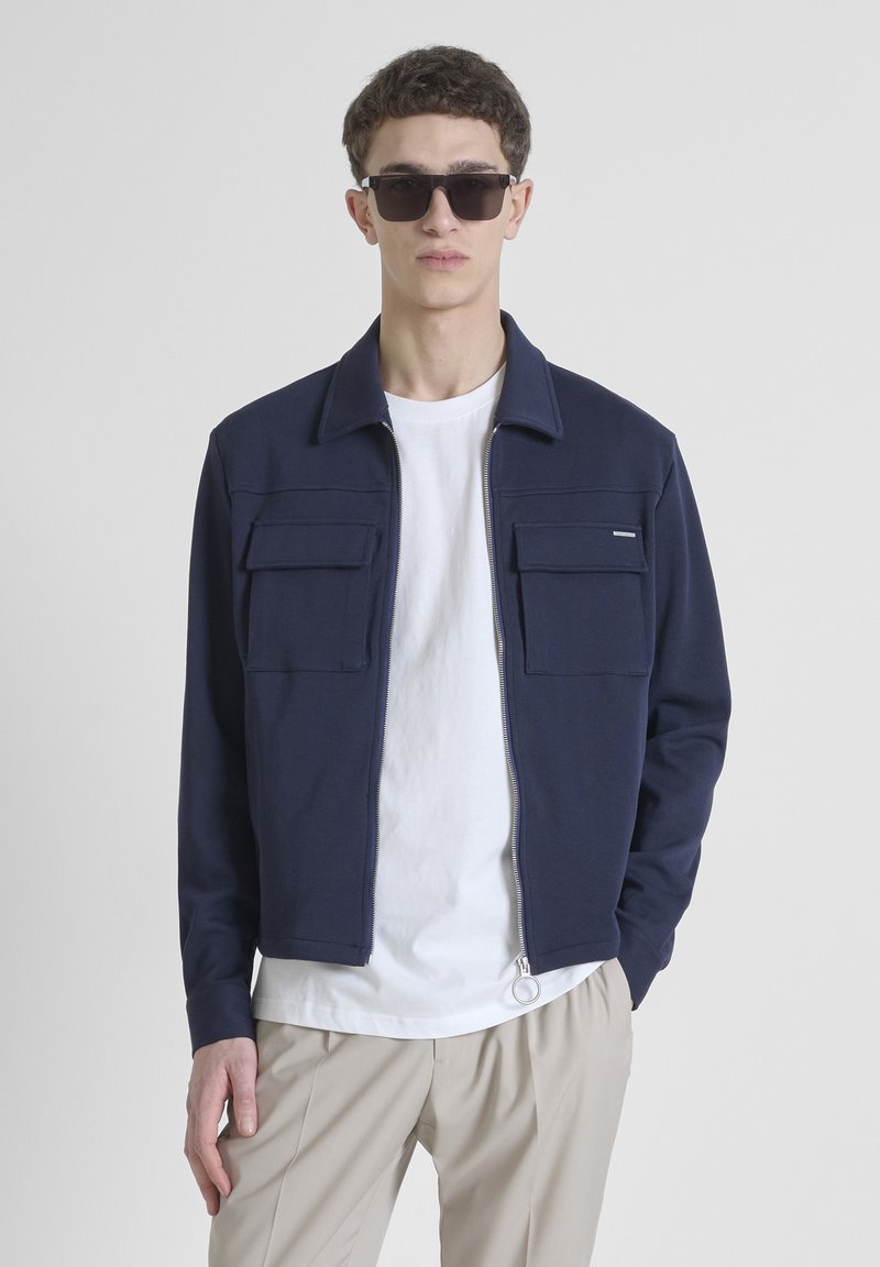 Marineblauwe jas met twee borstzakken, ritssluiting aan de voorkant en een kraag. Draagt over een wit T-shirt en beigekleurige broek. Textuurstof, casual pasvorm.