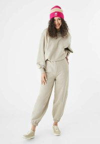Un sweat-shirt oversize beige et un pantalon de jogging assorti avec des poignets élastiques, associés à des baskets beige et à un bonnet en tricot rayé rose avec un logo.