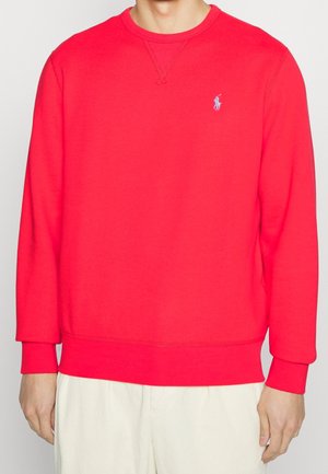 Mand iført en knaldrød langærmet sweatshirt med en lille lyseblå broderet logo på venstre bryst, kombineret med lyse bukser.