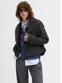 Temno karirano dvoredno plašč z velikimi reverji, noseno čez modro jeans srajco in črtasto srajco, v kombinaciji s sivimi hlačami in sončnimi očali.